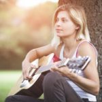 young woman playing acoustic guitar leaning on a tree