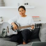 Woman in white long Sleeves on a couch playing the guitar