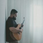 man in a room sunlit through curtains leaning back against a wall playing guitar