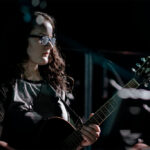 bespectacled woman in black playing guitar