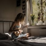 young woman playing guitar on her bed with flowers in the window