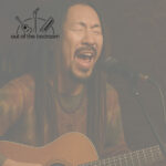 an early 40s hippie-looking man with dreadlocks in an open mic in a basement bar playing acoustic guitar and singing passionately into a microphone