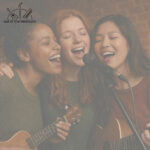 Three young women singing around a microphone at an open mic night