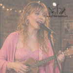 a young woman dressed in mostly pink clothing in an open mic in a basement bar singing like Taylor Swift into a microphone and playing ukulele